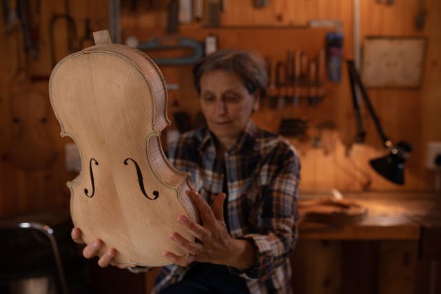 Front view of a senior Caucasian female luthier sitting and working on a violin at a workbench in her workshop, inspecting her handiwork and holding the unfinished violin body, with tools hanging up on the wall in the background.