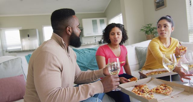 Three diverse male and female friends eating pizza and having wine in living room in slow motion. Friendship, leisure and lifestyle concept.
