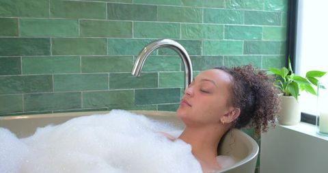 African american woman soaking in bubble bath with green tiles, chrome faucet, sunlight