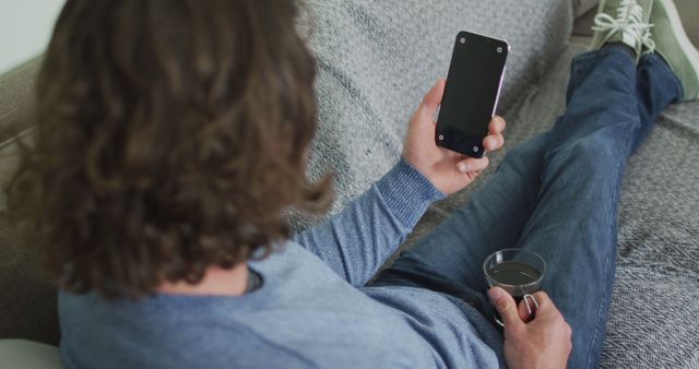 A man with long hair sitting casually on a couch, holding a smartphone and a transparent drink cup. Suitable for use in contexts involving relaxation, mobile technology, staying at home, casual living, and personal leisure time.