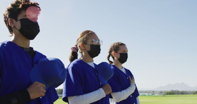 Three teen softball players standing in a row on a field, holding blue caps over their hearts and wearing blue uniforms with black face masks. Useful for themes related to health and safety in sports, teamwork, and efficacy. Ideal for articles, blogs, or advertisements promoting youth sports, COVID-19 safety measures, or teamwork and discipline.
