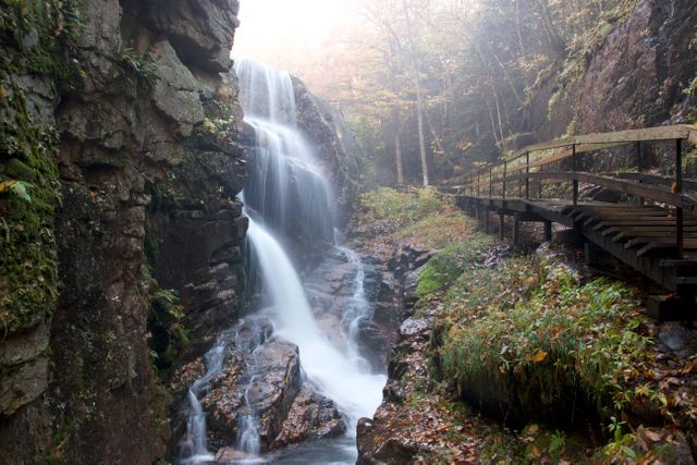 Majestic waterfall flowing through a rocky forest gorge during autumn season. Wooden bridge runs alongside the waterfall. Mist creates an ethereal atmosphere. Perfect for nature, outdoor travel, adventure, and serenity themes.