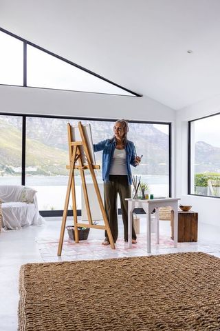 Middle-aged female artist painting at wooden easel in modern seaside studio with bay view