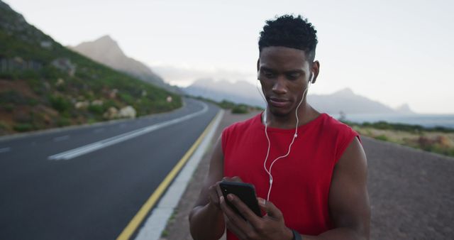 A fit young man is using a smartphone and headphones while standing on an empty road in a scenic natural setting. He is focused on selecting music or tracking his workout activity. This image can be used for themes related to fitness, technology, music, outdoor activities, health, and wellness. It is ideal for fitness apps, tech products, health blogs, and motivational promotions.