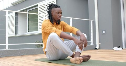 Young African American man sitting on green yoga mat on modern balcony in mustard top