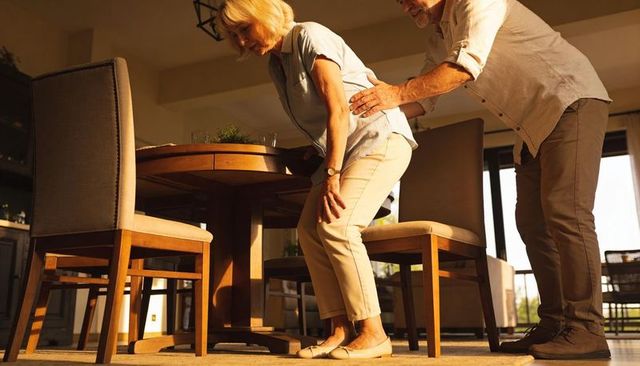 Senior couple at round dining table, man steadying woman rising from chair in sunlit home