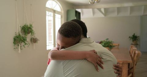 Father and son sharing warm embrace in sunlit dining room with hanging plants