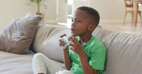 African-american boy playing video game on modern sofa holding wireless controller