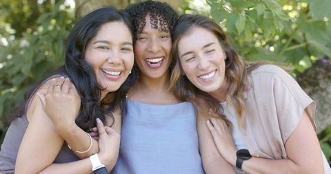 Laughing diverse women linking arms in sunlit garden, friendship and unity outdoors