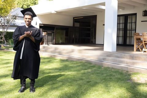 Graduate celebrating on backyard lawn wearing cap and gown, clapping in sunshine