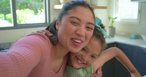 Mother and daughter taking selfie in sunlit kitchen laughing and hugging