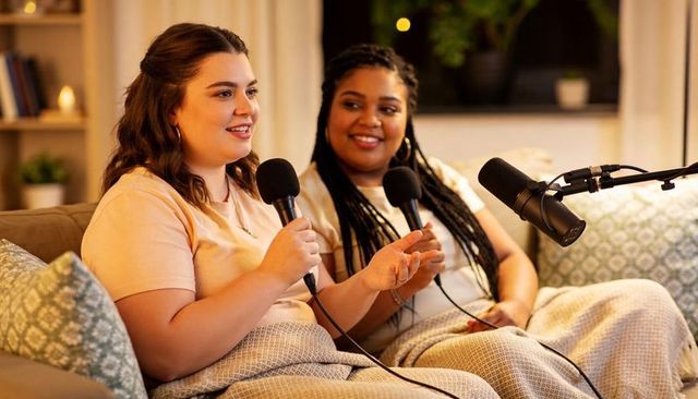 Two women recording cozy home podcast on sofa with microphones and boom mic