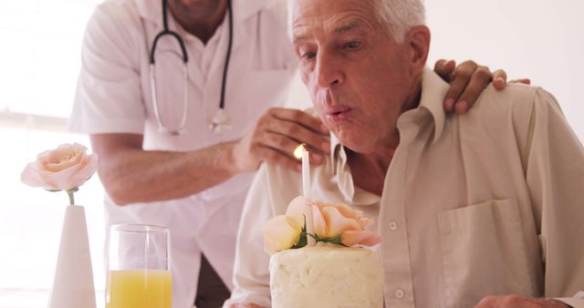 Elderly man celebrating birthday in a hospital setting with doctor assisting him. He is blowing out a candle on a small birthday cake with flowers. A vase with a rose and a glass of orange juice are on the table. Ideal for topics on elderly care, senior health, celebrations in healthcare settings, and compassionate medical care.