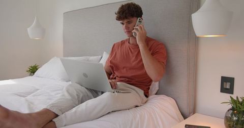 Working from Bed Man Talking on Phone while Typing on Laptop in Minimal Modern Bedroom