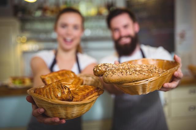 Two cafe employees, a waiter and waitress, smiling and holding baskets filled with fresh bread and pastries. Ideal for use in marketing materials for cafes, bakeries, or hospitality services, showcasing friendly service and fresh products.