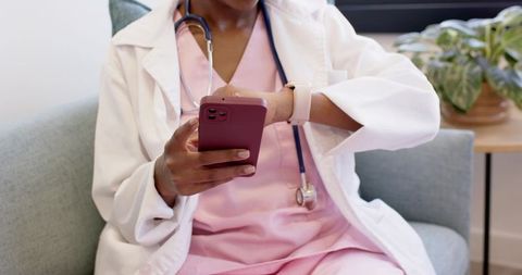 Nurse checking smartwatch while tapping smartphone, wearing pink scrubs and white coat