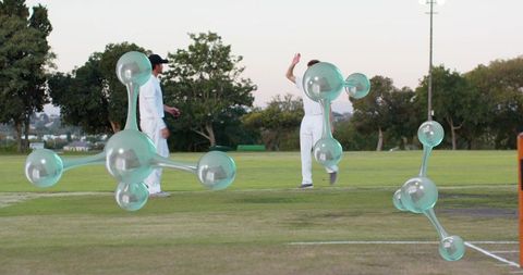 Cricketers celebrating at dusk with floating turquoise molecules over grass pitch