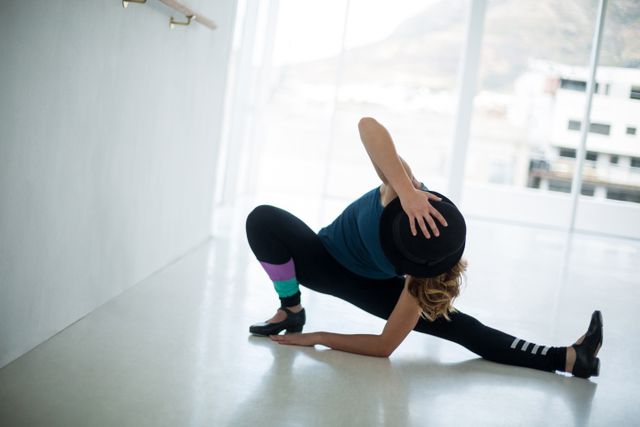 Dancer practicing in a bright studio, wearing a hat and performing a stretch. Ideal for use in articles about dance training, fitness routines, contemporary dance, and artistic performances. Can also be used in promotional materials for dance studios or fitness programs.