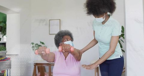 Senior woman exercising with caregiver wearing masks using pink dumbbells for rehabilitation