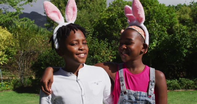 Two African American children are embracing and smiling while wearing bunny ears in an outdoor park setting. Arty grass and trees, indicating a natural, sunny day in the background. Great for themes concerning family, childhood, outdoor activities, celebrations, and festive events such as Easter.