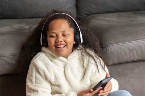 Smiling girl listening to music on headphones holding smartphone on living room floor