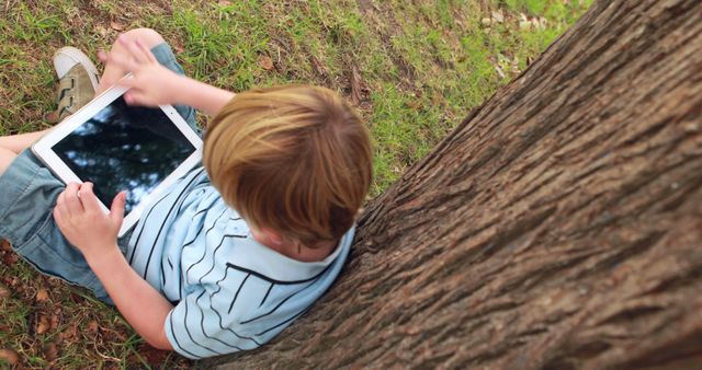 Young boy leaning against tree using a tablet in a park. Ideal for depicting childhood in digital age, outdoor learning, leisure activity, technology’s role in education, and promoting outdoor family time.