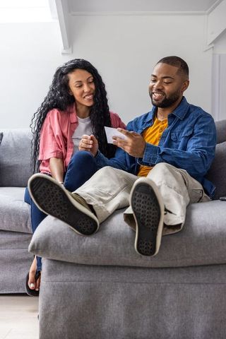 Diverse couple sitting on sofa holding printed card and smiling, casual home moment