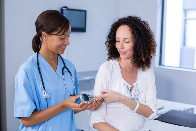 This image shows a nurse checking a pregnant woman's blood sugar level in a clinical setting. The nurse is using a glucose meter while the patient is smiling, indicating a positive interaction. This image can be used for healthcare websites, prenatal care articles, medical brochures, and educational materials about pregnancy and diabetes management.