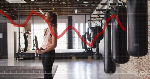 Female athlete holding jump rope in gym with punching bags and red performance graph