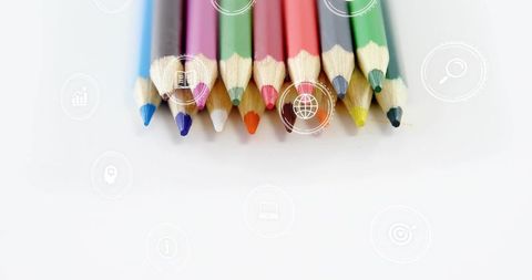 Color pencil tips aligned on white surface showing faint digital icons for creativity