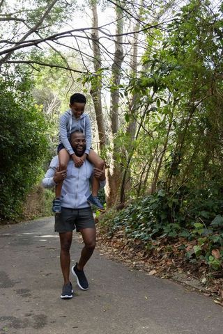 African american father giving son shoulder ride on woodland trail, smiling family walk