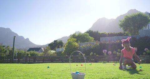 African american girl collecting easter eggs in sunlit backyard wearing pink bunny ears