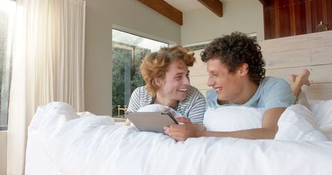 Cheerful Gay Couple Relaxing with Tablet in Cozy Bedroom