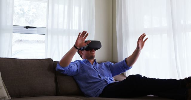 Man wearing VR headset while sitting on sofa, interacting with virtual environment. Ideal for use in articles about virtual reality technology, home entertainment, modern technology trends, gaming experiences, and digital leisure activities.
