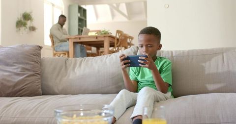 African american boy using smartphone while father working on laptop in sunlit living room