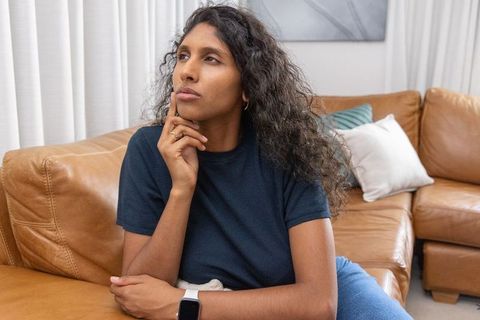 Contemplating african american woman sitting on leather sofa wearing smartwatch cozy living room