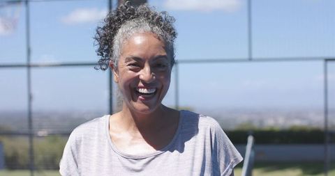 Smiling middle-aged woman standing on outdoor basketball court wearing light tee in bright sunlight