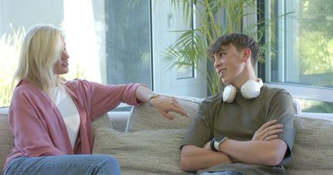 Woman talking with teenage boy on sofa with headphones and smartwatch in bright living room