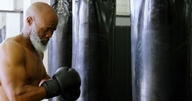 Elderly man wearing boxing gloves trains with punching bags in gym. Perfect for fitness, health, or active lifestyle promotions, showcasing senior fitness, or illustrating articles on staying fit in older age.