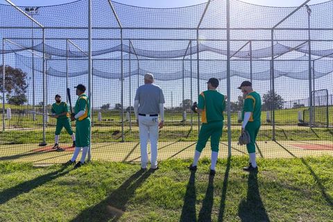 Coach directing baseball batting practice in outdoor netted cage with green-uniform players