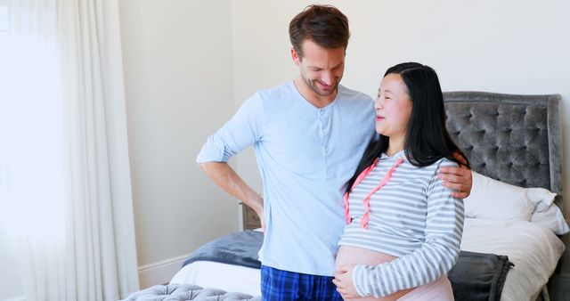 Happy diverse husband and pregnant wife in nightclothes embracing in sunny bedroom, copy space. Couple, communication, pregnancy, preparation, relationship, togetherness and domestic life, unaltered.