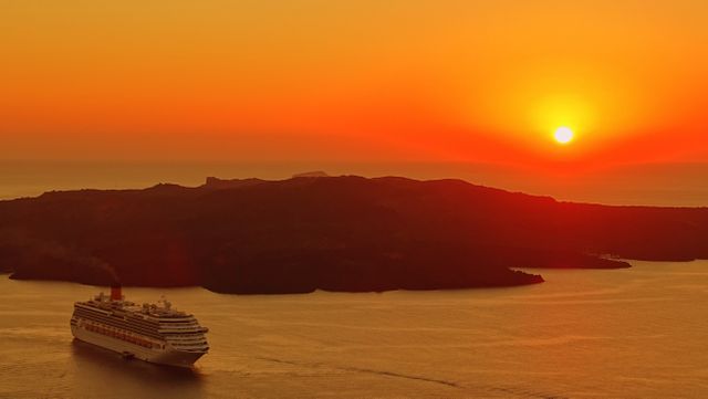 Cruise ship sailing past an island during sunset with an orange sky and tranquil waters. Perfect for travel brochures, tourism advertisements, and vacation planning materials.