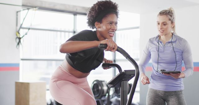Image of diverse female fitness trainer and woman on exercise bike working out at a gym. Exercise, fitness and healthy lifestyle.