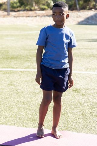 African american boy standing barefoot on pink mat on sunny sports field practicing yoga