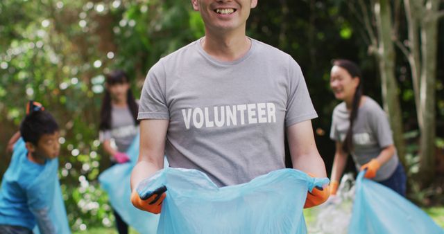 Smiling asian man wearing volunteer t shirt holding refuse sack for collecting plastic waste. eco conservation volunteers doing countryside clean-up.