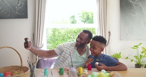African american father and son taking selfie while painting colorful easter eggs together