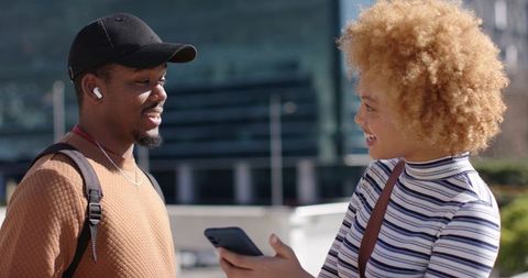 Smiling friends chatting on urban plaza, woman holding smartphone, man with backpack