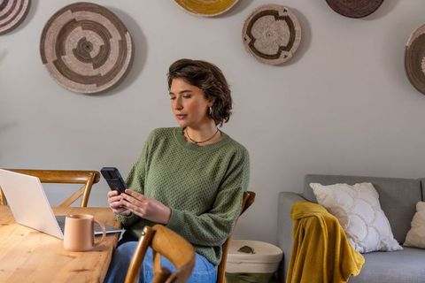 Adult woman working remotely at dining table, checking phone beside laptop with coffee