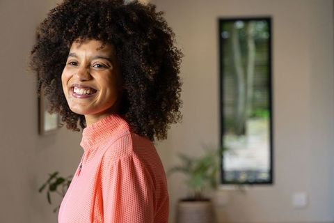 Smiling African American woman wearing coral zip-up top with full curly hair in cozy home