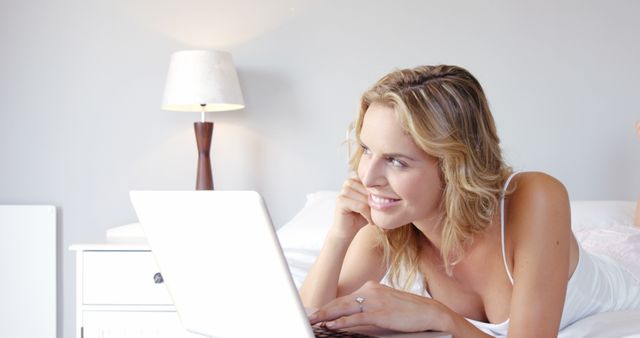 A smiling woman is using her laptop while lying in bed in a cozy bedroom with soft lighting. She appears to be relaxed and content, creating a serene home setting atmosphere. This image can be used to depict modern lifestyle, comfort, and technology usage in home environments. Ideal for blog posts, website banners, advertisements for home decor, technology products, or relaxation-themed content.